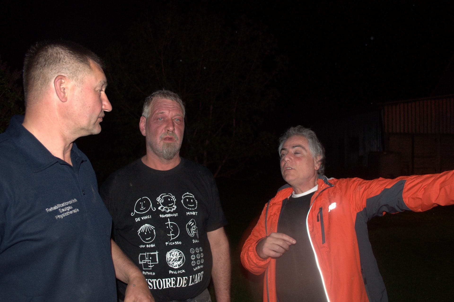 Three individuals are standing outside at night, with one person pointing to the right.