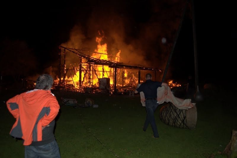 NIKON CORPORATION NIKON D5000 · 18 mm · f/3.5 · 1/200 s A nighttime outdoor scene with a large fire burning in the background, consuming a structure. A person in a red jacket is standing in the foreground, and another person is holding a large object, possibly a barrel, in the background.