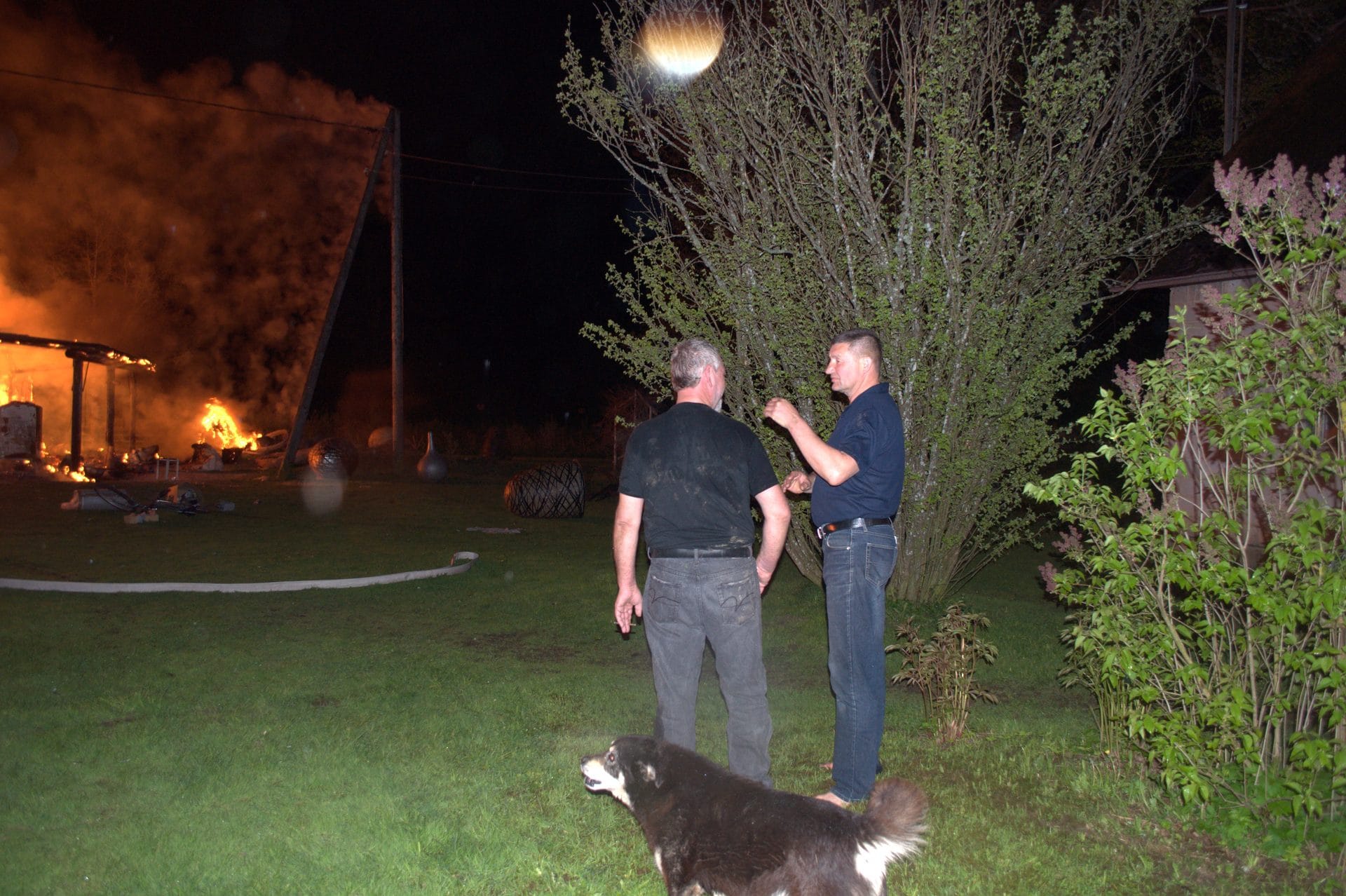 Two men and a dog are standing in a yard at night, with a large fire in the background.