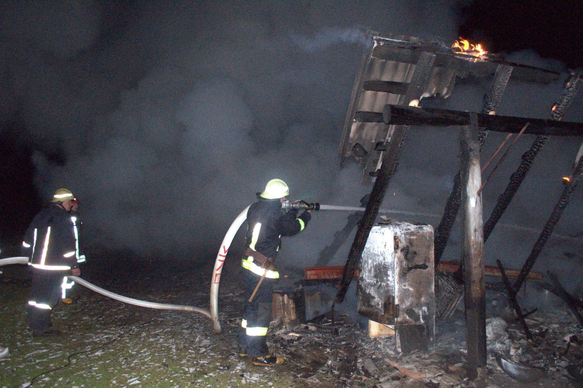 Two firefighters are battling a fire at night, using a hose to spray water on the flames.