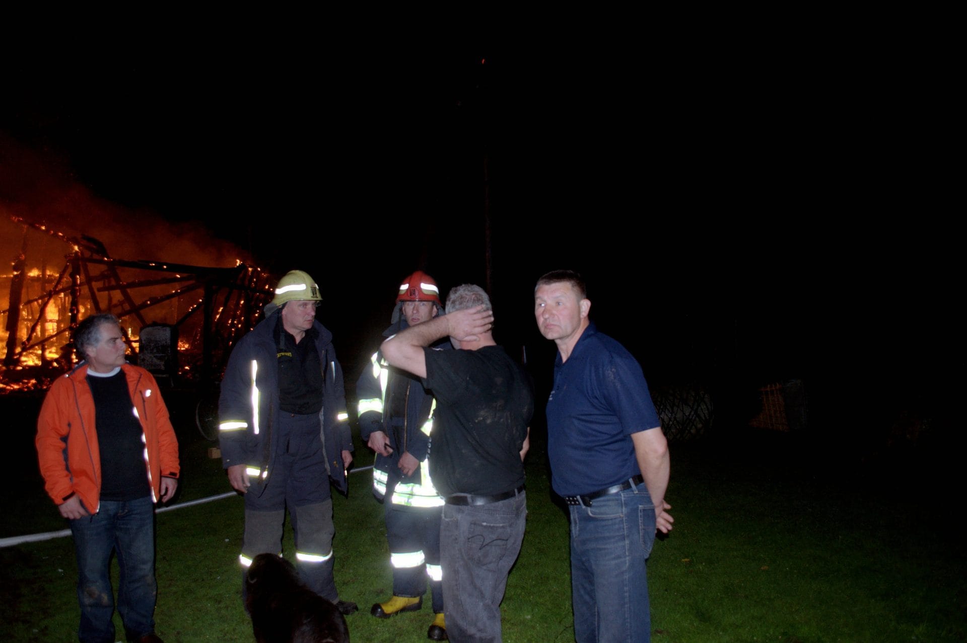 NIKON CORPORATION NIKON D5000 · 18 mm · f/3.5 · 1/60 s A group of people are gathered outside at night near a fire. The fire is burning a wooden structure. The people are wearing reflective clothing and helmets, indicating they might be emergency responders. One person is wearing a blue shirt and jeans, and appears to be a civilian. A dog is visible in the foreground.